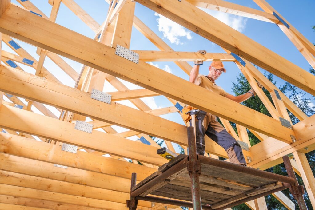 A carpenter carefully constructs the wooden frame of a house in a bright, sunny environment, showcasing detailed work and craftsmanship in progress.
