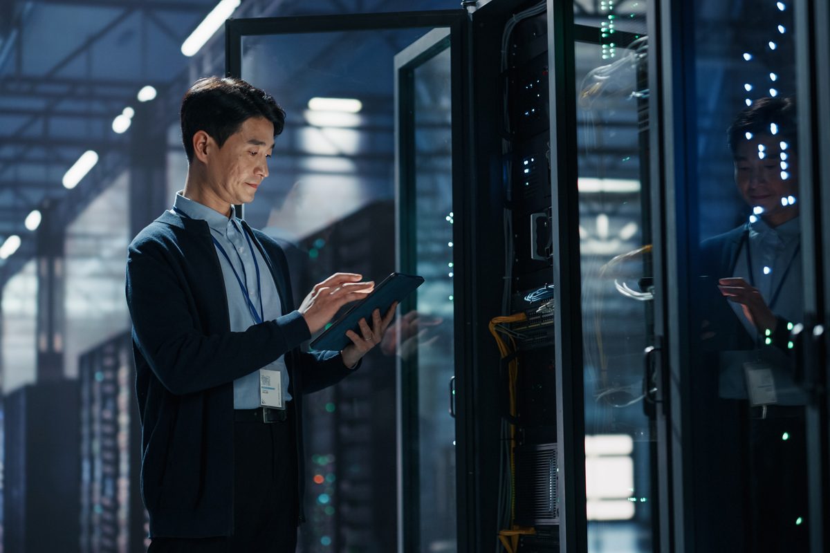 Asian Male It Specialist Using Tablet Computer to Configure Network Settings Beside Open Server Rack, Managing Data Migration and Uptime Analytics in AI Cloud Computing Colocation Facility