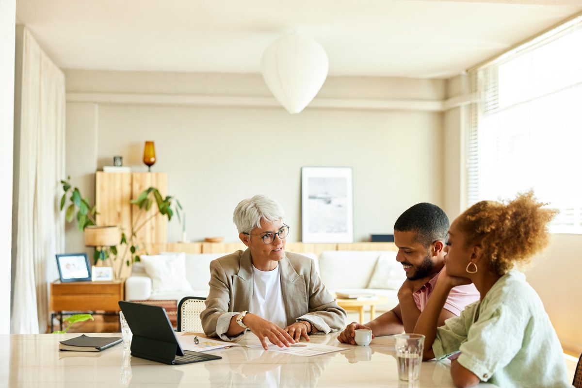 Mature female advisor discussing with couple in apartment. Man and woman are listening to finance manager. They are sitting at table.