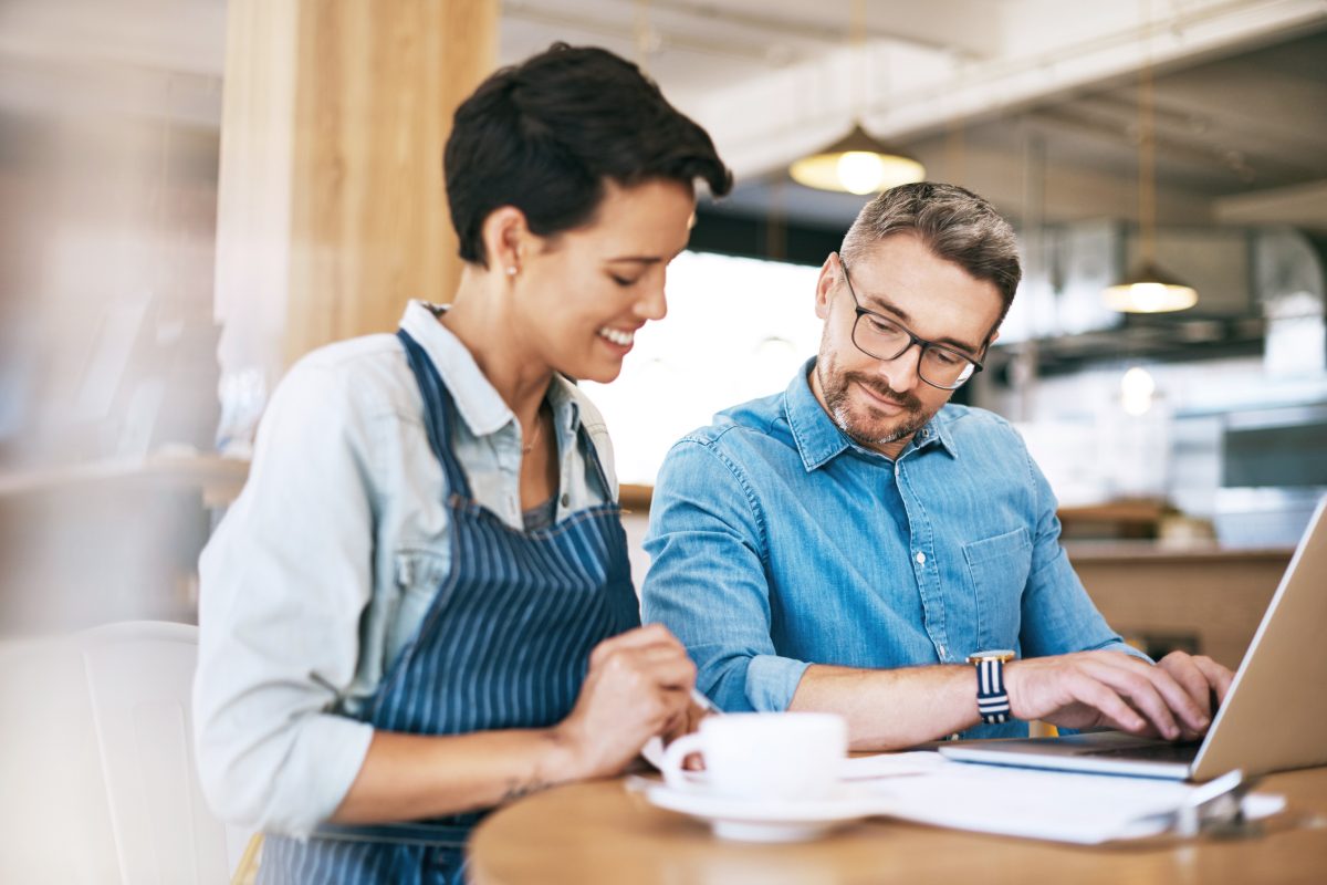 Shot of a man and woman going through paperwork and using a laptop together in their coffee shop