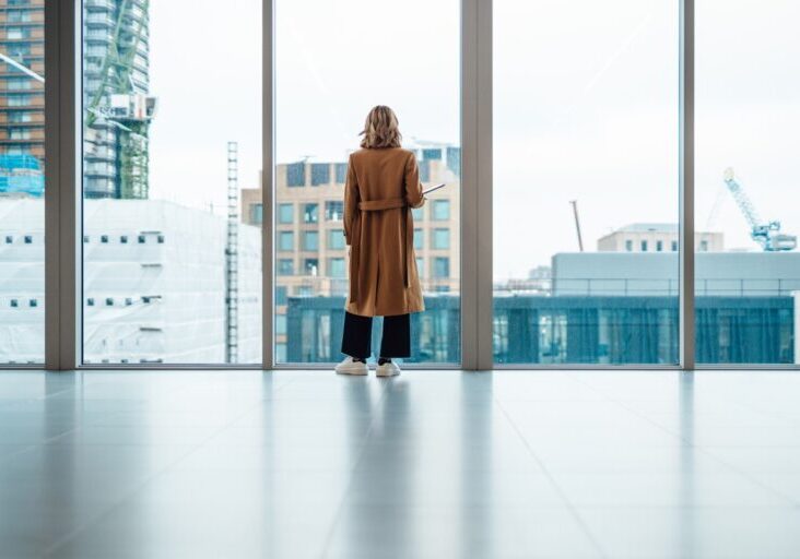Back view of businesswoman standing and looking through window in conference room. She is thinking business strategy and making new business plan. Holding a laptop on her arm. Transgender female pursuing her dreams and successful career.