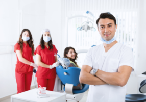 Dental-team-one-male-dentist-in-white-and-2-assistance-in-red-next-to-a-smiling-patient-in-a-blue-chair
