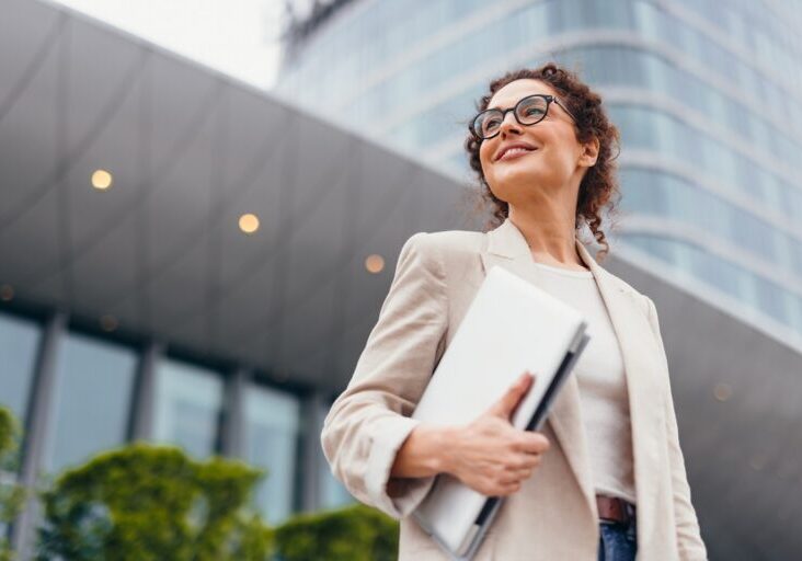 Happy businesswoman in eyeglasses holding a laptop, smiling confidently while looking away, standing near a modern office building on a bustling city street