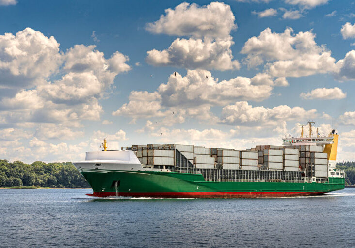 A full loaded cargo ship sets out to sea. Coastal landscape background. Sunny day, blue sky with clouds.