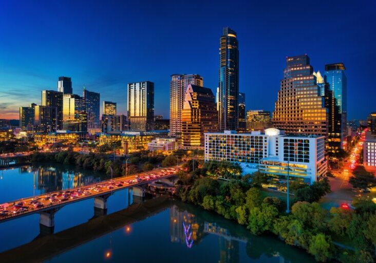 Downtown Austin skyline, elevated view, at dusk with the sunset reflections on the building facade, and the Lady Bird Lake / Town Lake / Colorado River with heavy traffic on the bridges in the foreground.