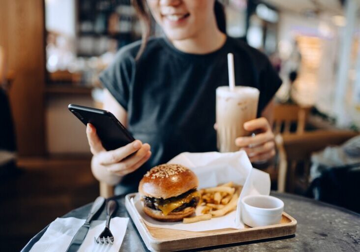 Cropped shot of smiling young Asian woman using smartphone while enjoying her lunch, cheeseburger with french fries and iced coffee in a stylish cafe. People, food, lifestyle and technology concept
