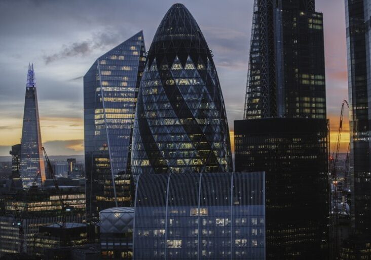 Captivating view of London’s financial district during twilight, featuring iconic skyscrapers like The Gherkin and The Shard. The blend of warm sunset hues and artificial building lights highlights the modern architecture and vibrant city life, showcasing the heart of London’s business hub