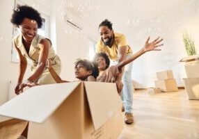 Playful African American parents having fun while pushing their small kids in carboard box at new apartment. Copy space.