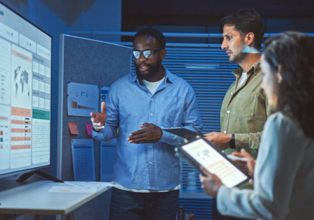 In a blue lit room, a team of developers stand around a television screen displaying charts and graphs